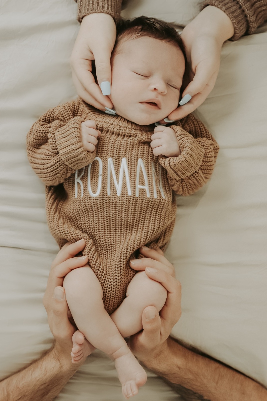 Sleeping newborn Roman in a brown knitted outfit cradled by his parents’ hands