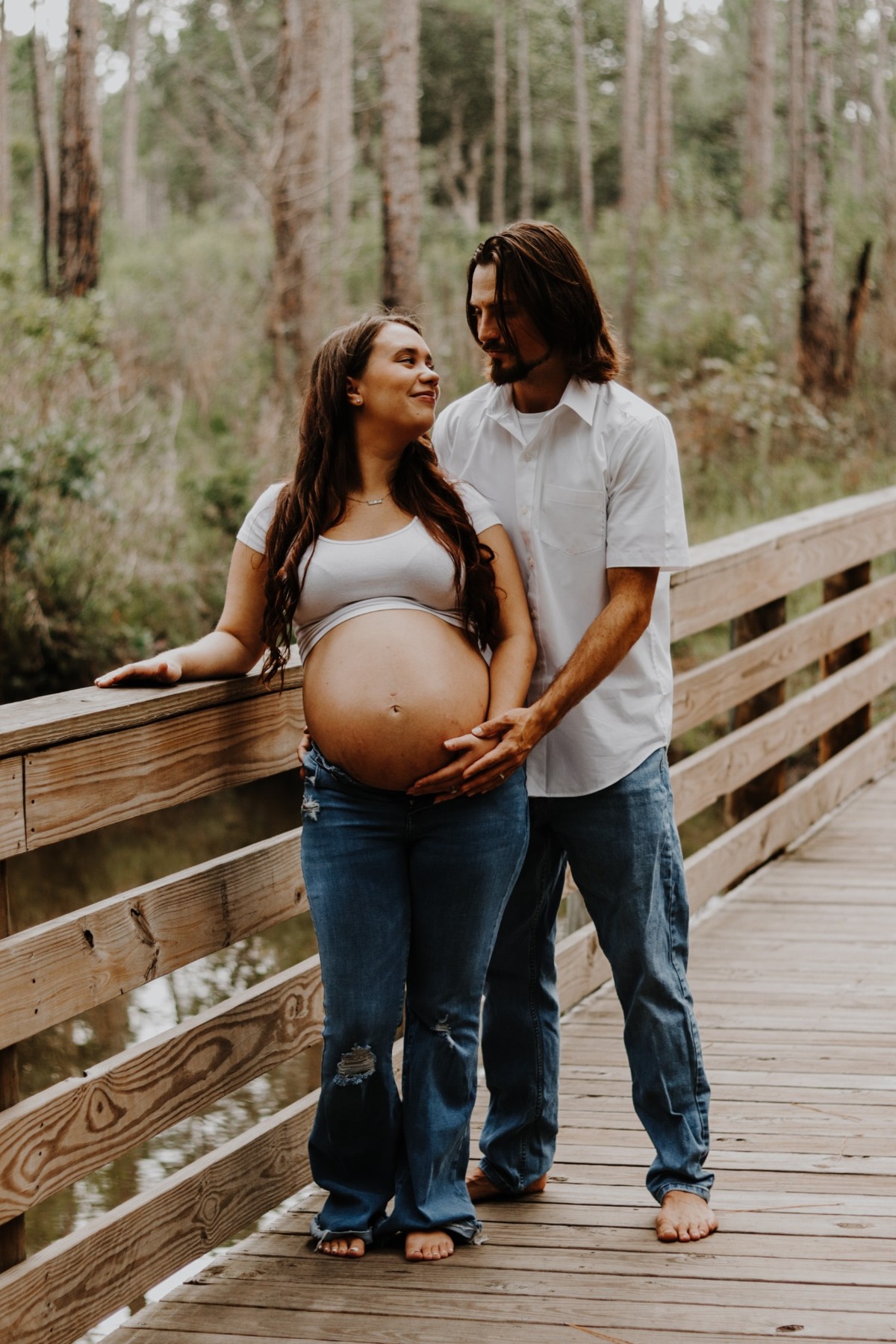 Expectant parents sharing a smile on a wooded boardwalk